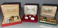 Three open boxes each containing vintage men's jewelry including cuff links and tie clips. Left box red with satin lining labeled 'Finest Quality JEWELLERY FOR MEN' holds gold-tone items. Center red velvet and satin box labeled 'Century' holds silver-tone rhinestone cuff links. Right green and brown box holds green-accented cuff links.