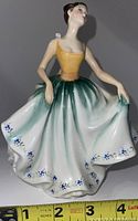 Front view of Royal Doulton porcelain figurine of a young woman in a dress holding the skirt out with her hands, showing the detailed floral decoration and color transition on the skirt and bodice.