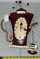 Front view of the teapot shaped wall clock showing red body with black circular patterns, gold handle and spout, cream scalloped base, oval clock face and attached steaming cup.
