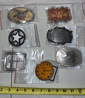 Overview of eight assorted vintage belt buckles in plastic bags on a white surface with measurement scale, varying designs including eagle, star, clock, and cowboy boot.