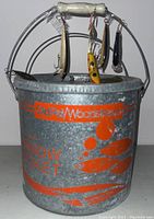 Photo of vintage galvanized metal minnow bucket with orange branding and several fishing lures hanging from handle.