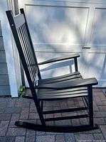 Side profile of black wooden rocking chair showing vertical slats on seat and back, curved rockers, flat armrests.