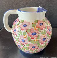Side view of the round ceramic pitcher decorated with colorful hand-painted flowers and a blue-accented rim and spout.