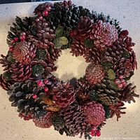 Top-down view of a 12.5 inch diameter Christmas wreath made of painted and natural pinecones with red berries and green leaves on a white countertop.