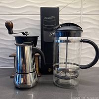 Two French coffee presses and a stainless steel single espresso maker with wooden-handled manual coffee grinder behind