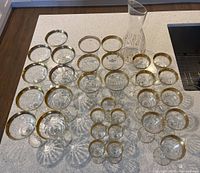 Set of crystal glasses arranged on countertop including wine, sherry, liqueur, and champagne glasses with gold rims alongside a clear glass carafe with etched geometric design.