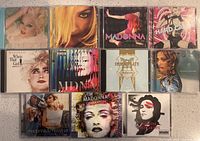 Photo showing 11 Madonna CD albums arranged in 3 rows on a white surface, featuring different album covers including studio, compilation, and collaboration albums.