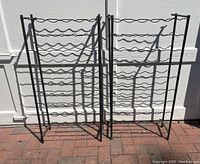 Photo of two black metal wire wine racks side by side showing vertical rectangular frame and horizontal wavy wire rows for bottles.