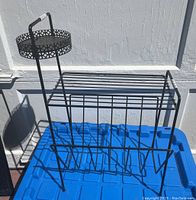 Front and side view of a black coated metal magazine rack with a round elevated compartment and multiple vertical angled slots for magazines.