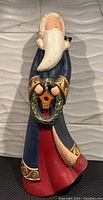 Front view of ceramic Santa figurine with long blue coat, white beard, holding a green wreath with small birdhouse detail.