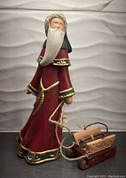 Photo of 10-inch Santa Claus ceramic figure wearing a red robe with gold trim, holding a rope attached to a sled carrying firewood.