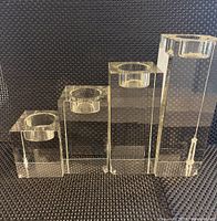 Four clear crystal tea light candle holders arranged by height on a textured black background. Reflective surfaces and transparency highlight the crystal quality.