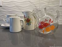 Three vintage pitchers and one cream and sugar set placed on a countertop with a background tile wall