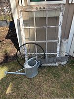Photo showing the antique wooden window frame, black metal wheel, galvanized watering can, 'WELCOME' metal sign, and stone-like pedestal on grass near a shed.