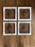 Top-down view of four square decorative wall hangings with distressed white wooden frames and ornate metal floral central designs placed on wooden floor.