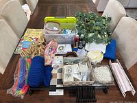 Full overview of all arts and crafts supplies on wooden table, including paints, brushes, pipe cleaners, rope, foliage, ribbons, yarn, beads, and more.