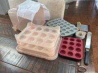 Photo showing three stacked cupcake trays in pink, gray, and red colors, a white apron, metal icing scraper, baking thermometer, icing spreader, and baking strips all arranged on a wooden surface.