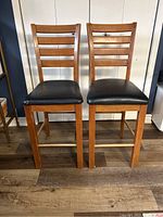 Front view of two wooden bar stools with black faux leather seats on wood floor.