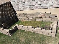 L-shaped arrangement of rectangular brick pavers stacked loosely on grass beside a house wall with a lattice fence on one side.