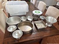 Photo showing multiple metal cake pans of various shapes and sizes arranged on a wooden table with a clear plastic tote in the background.