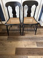 Front view of two identical antique cane-back chairs with black finish and natural cane seats on wooden floor
