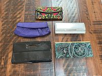 Set of five clutches arranged on wood floor: colorful floral embroidered fabric, purple pleated fabric, white textured with metallic bar, black vinyl with cut-out handle, blue paisley patterned with metallic frame.