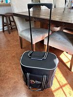 Front view of black cooler bag on wheels with handle extended, showing front pocket and brand logo.