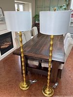 Two gold-toned floor lamps standing beside a wooden table, showing their full height and slender bases.