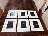 Top-down view of six white gallery wall frames laid out on a wooden table, each containing a black insert where the photos would go.