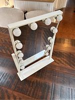 Front view of the white framed rectangular mirror with 12 round bulbs mounted around the perimeter, sitting on a white base stand on a wooden table.