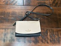 Front view of small two-tone Kate Spade crossbody purse with beige flap and black body showing the Kate Spade logo in gold-tone on flap.