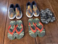 Top view of two pairs of slip-on canvas shoes with tropical leaf prints and two pairs of camouflage flip flops on wooden floor.