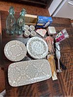 Full overview of kitchen items assembled on table including dinnerware, bottles, utensils, mixer, and accessories.