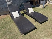 Two dark brown wicker chaise lounges on grass with one throw pillow on each.