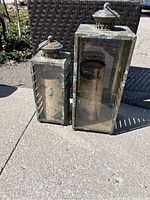 Front view of two antique metal outdoor lanterns with glass sides and candles inside, placed on pavement with dark brown wicker side table behind.