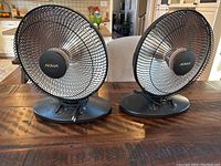 Two NOMA portable heaters side by side on wooden table showing front protective grills and bases.