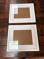 Two white rectangular wall hanging frames stacked on a wooden table, showing front with glass and matting.