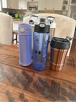 All five bottles and mug shown on table: purple insulated bottle, blue Under Armour bottle, two transparent Amino bottles with white lids, copper travel mug