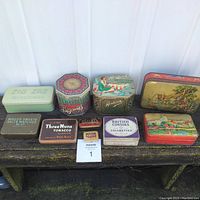 Front view of the 10 vintage tins arranged in two rows on a wooden bench. Shows a variety of colors, sizes, and designs including tobacco, medical, and accessories tins.