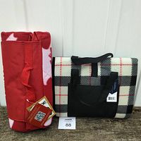 Two folded picnic blankets side by side, showing one red patterned and one black, white, red plaid blanket with carrying straps