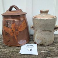 Front view of two pottery canisters side by side on wooden surface, showcasing dragonfly motif on brown canister and beige speckled canister with handles.