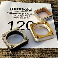 Three square rings placed on auction paper, showing the white ceramic ring with gold-tone screws and two stainless steel rings with screw motifs.