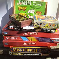 Photo showing stacked board games including 3 Scrabble sets, Word Mastermind, Kings Cribbage, Chinese Checkers, Farm Spelling Pictures puzzle and travel magnetic chess set on wooden surface.
