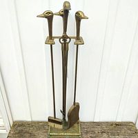 Full view of brass fireplace tool set on stand with duck head handles showing four tools: poker, brush, shovel, and tongs in brass with patina mostly on stand base.