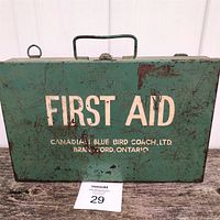 Front view of a green metal first aid kit box showing the 'FIRST AID' text and 'Canadian Blue Bird Coach Ltd, Brantford Ontario' printing with some rust and scratches.