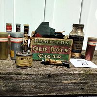 Photo showing wooden crate with old metallic powder dye containers including 'Sheffield Ready Mixed Gold Paint' glass jar and plastic vial '26 Venus Extra Brilliant Fire' along with other vintage tins and tubes.