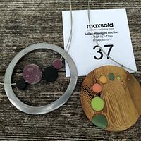 Two wire necklaces laid flat showing one metal circular pendant with attached wood beads and one wooden oval pendant with color inlays.