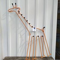 Full view of tall giraffe-shaped orange metal wall hanger with multiple black-tipped hooks along neck and legs, hung on white vertical panels.
