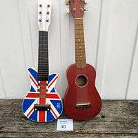 Front view of two stringed instruments: a small guitar painted with the Union Jack on the left and a reddish-brown ukulele on the right, both resting against a wall.