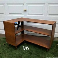 Front angled view of the plywood corner shelf showing its structure, two shelves, and brass capped feet on a grass background in front of a garage door.